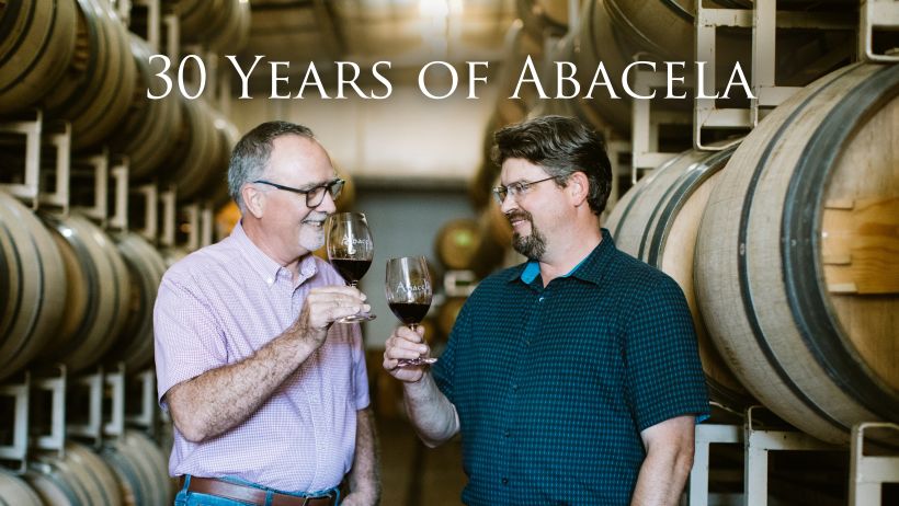 Abacela 30th anniversary image with Greg Jones and Andrew Wenzl holding a glass of wine in a barrel room
